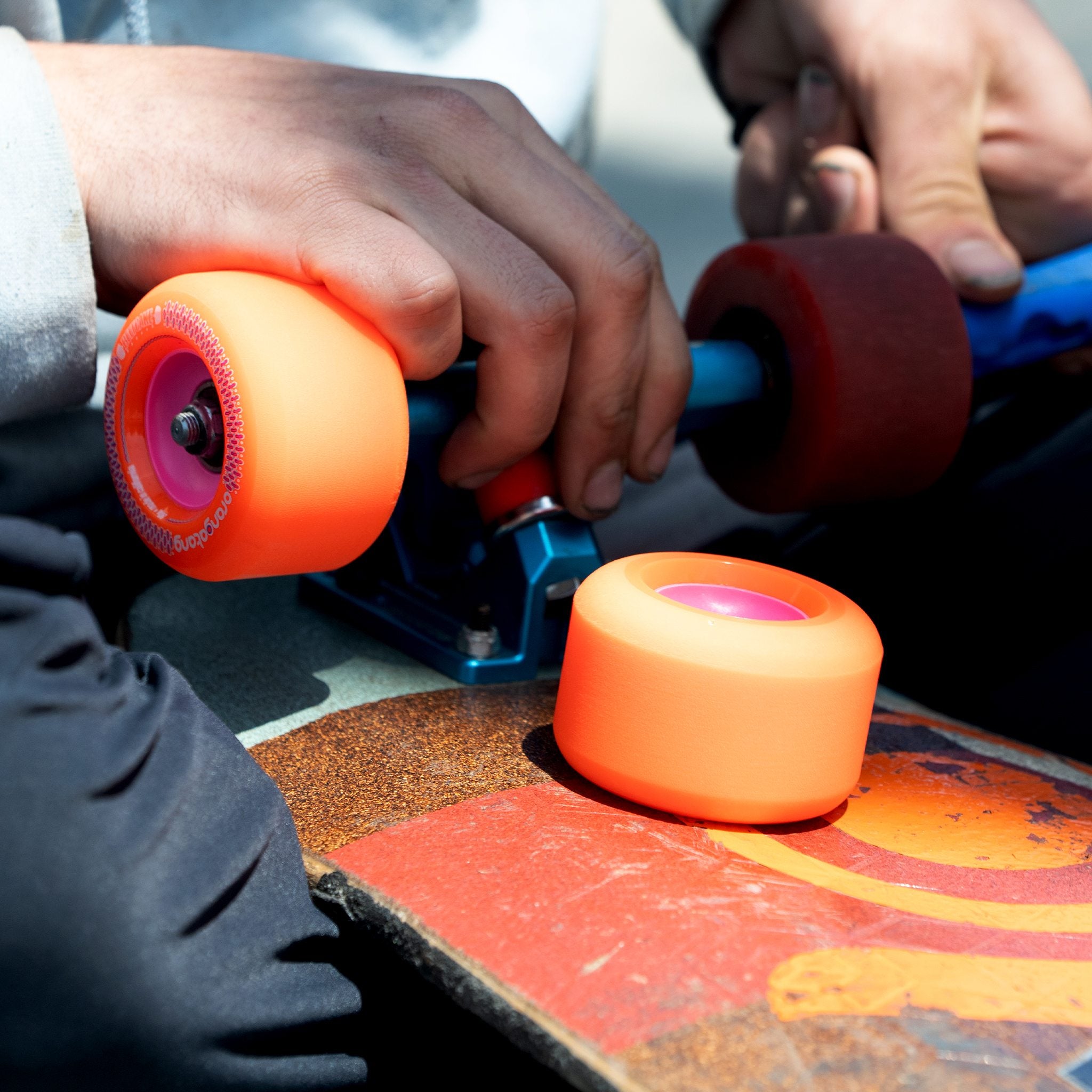 Skater swapping orange Orangatang Beefcake 73mm wheels onto blue skateboard trucks, with a loose wheel resting on the deck