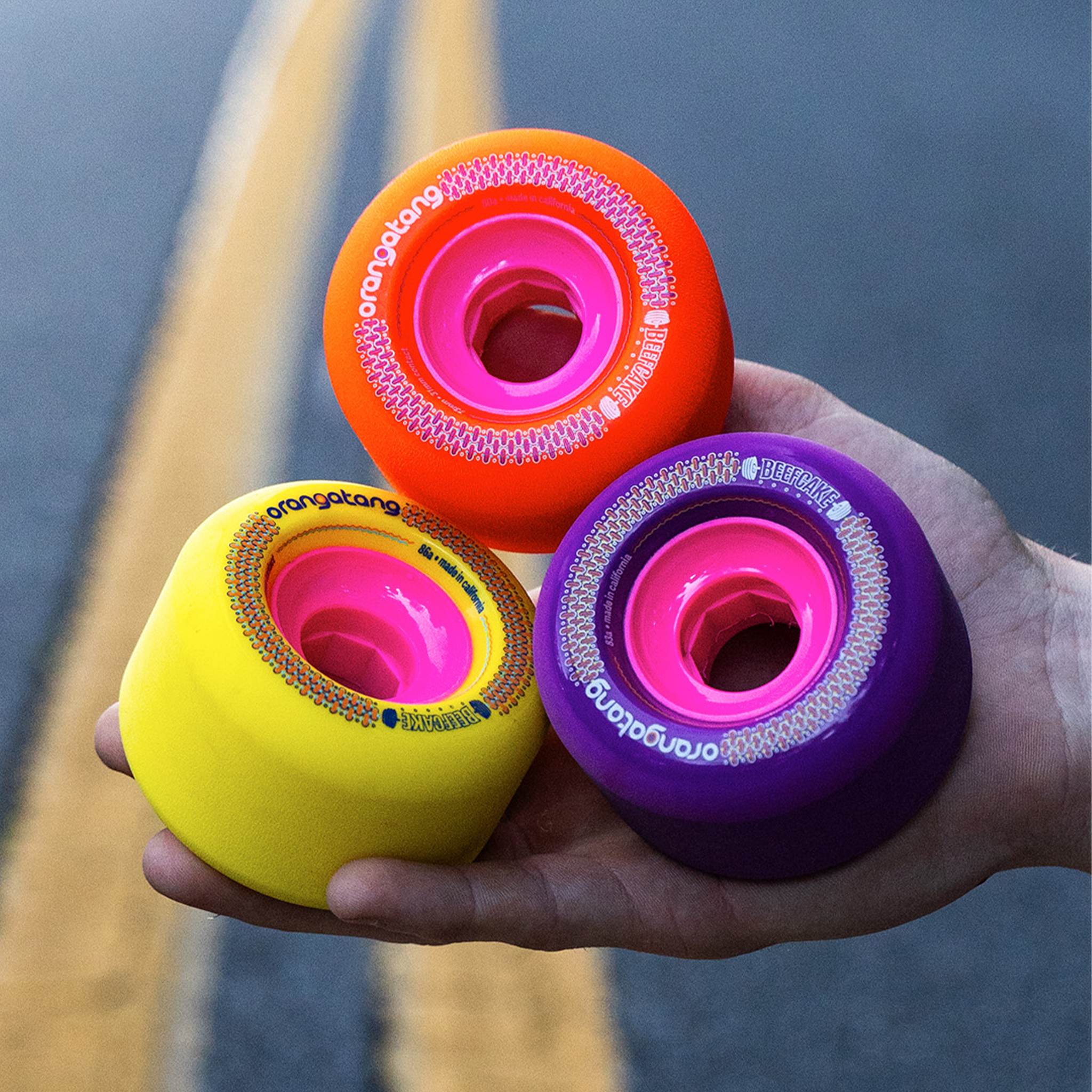 Hand holding three Orangatang Beefcake 73mm wheels in orange, yellow, and purple with pink cores over a road's yellow center line