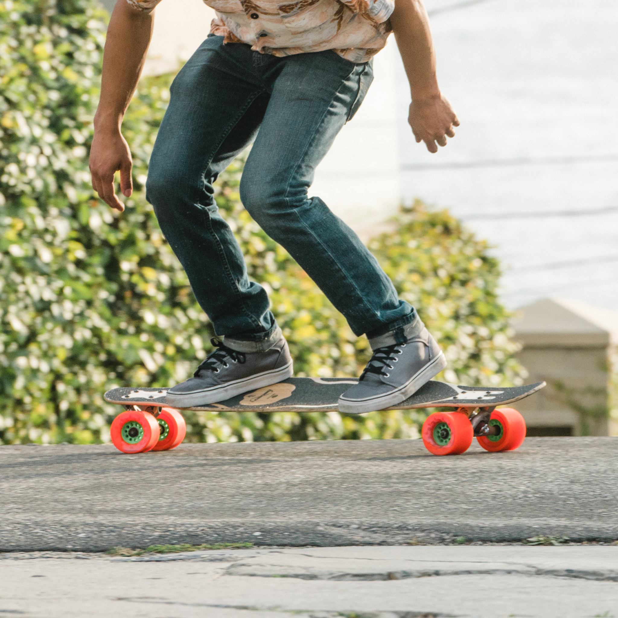 Rider in jeans and sneakers mid-pump on a longboard with orange Orangatang Caguama 85mm wheels along a suburban street
