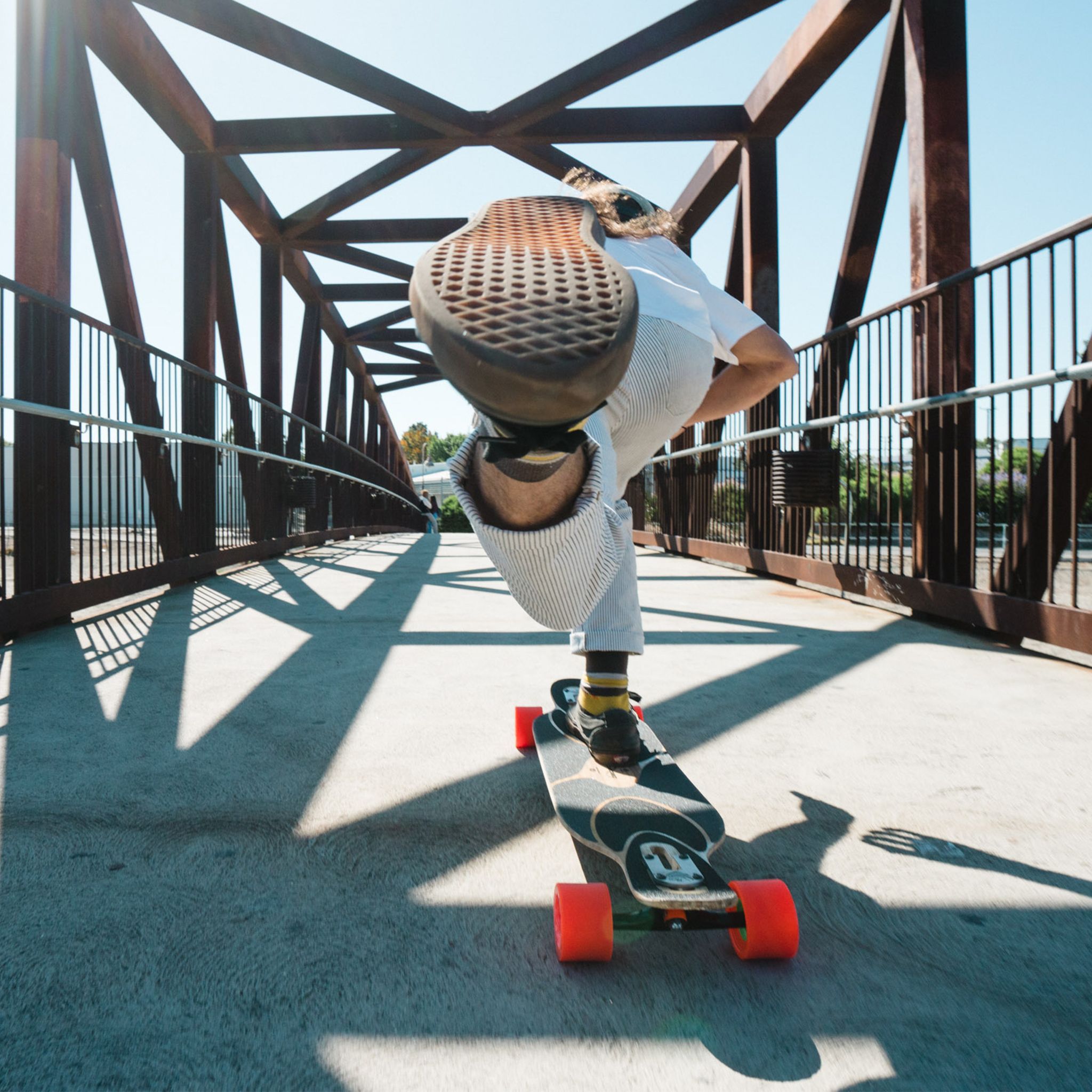 Low-angle view of a rider pushing a longboard across a steel truss footbridge on orange Orangatang Caguama 85mm wheels