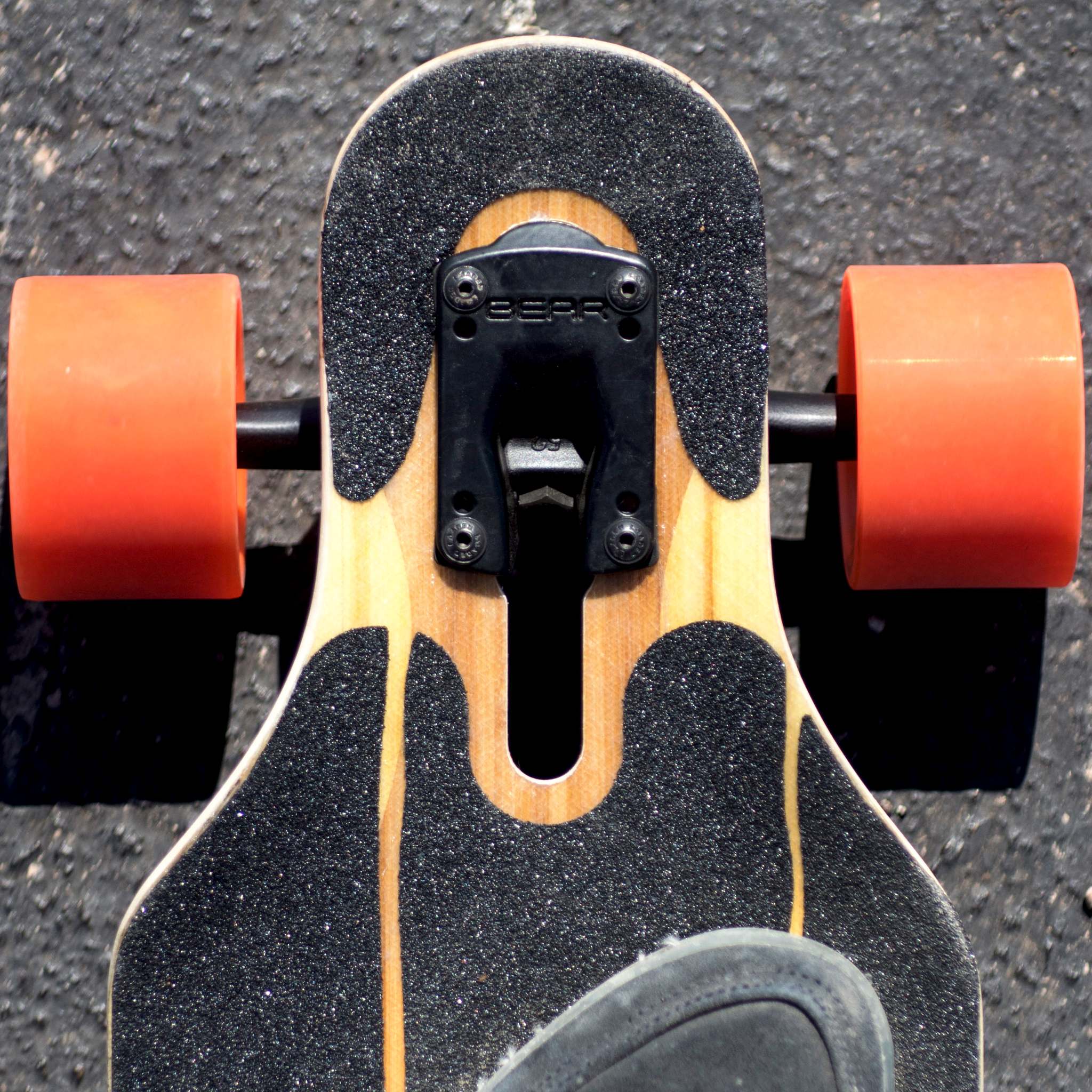Overhead view of a longboard's front truck with orange Orangatang Caguama 85mm wheels on asphalt, rider's shoe in frame