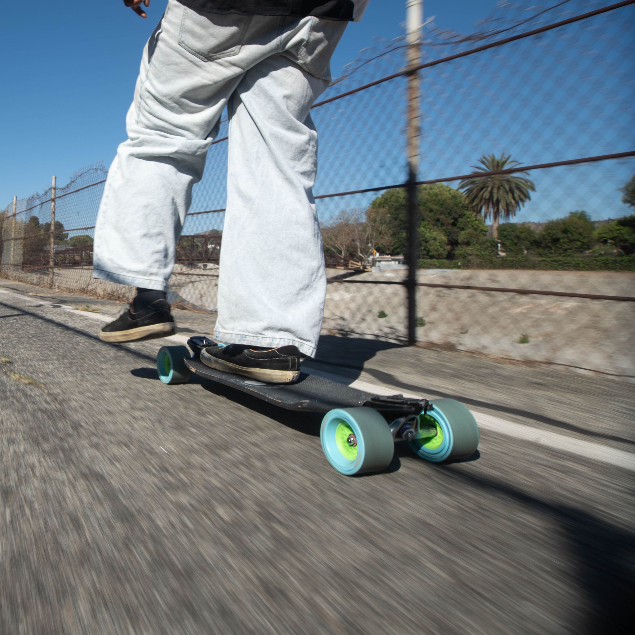Rider in wide-leg jeans cruising a longboard on light blue Orangatang Dad Bod 105mm wheels along a fenced path