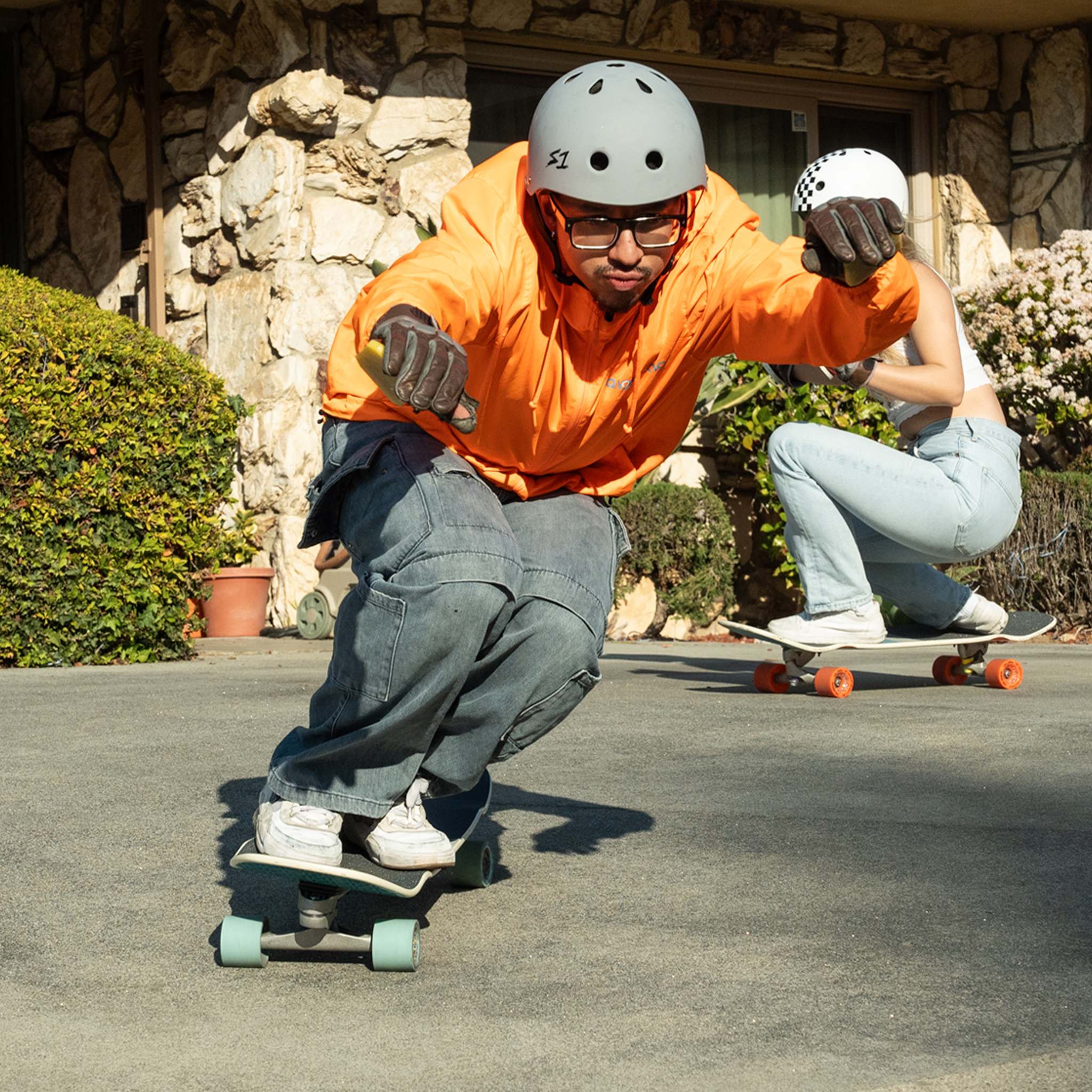 Two skaters in helmets crouched low, carving cruiser boards with Orangatang Love Handles 65mm wheels past a stone building