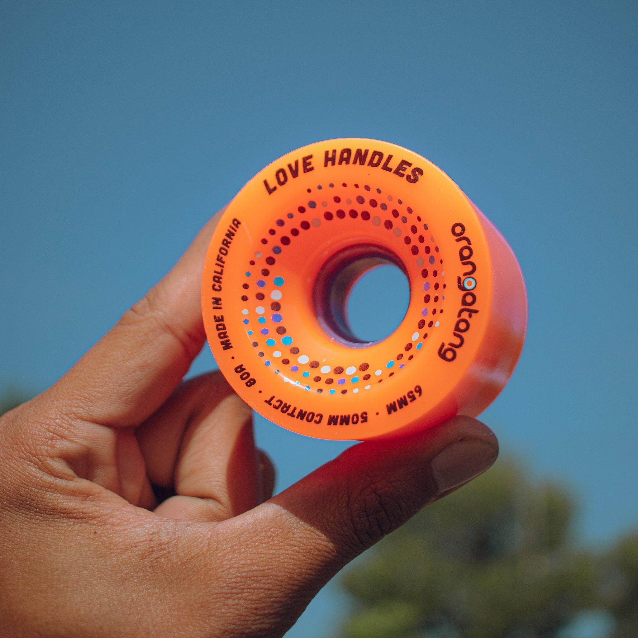 Hand holding up an orange Orangatang Love Handles 65mm 80a wheel against a clear blue sky