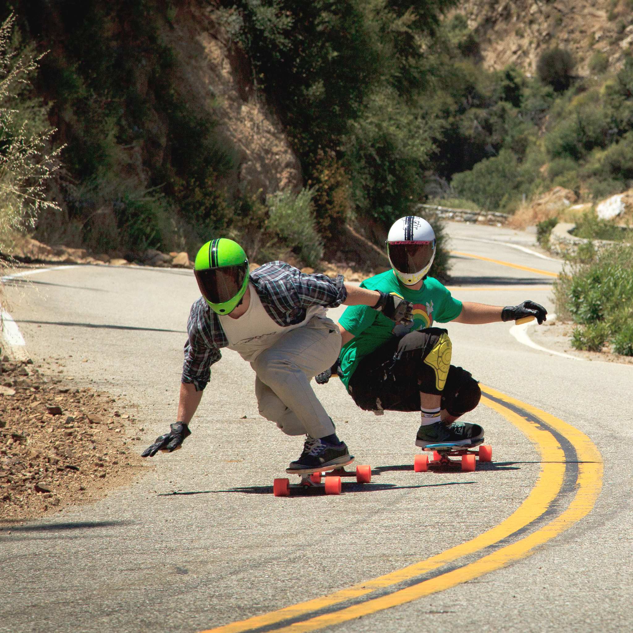 Two downhill skaters in full-face helmets carving a mountain road turn on orange Orangatang In Heat 75mm wheels