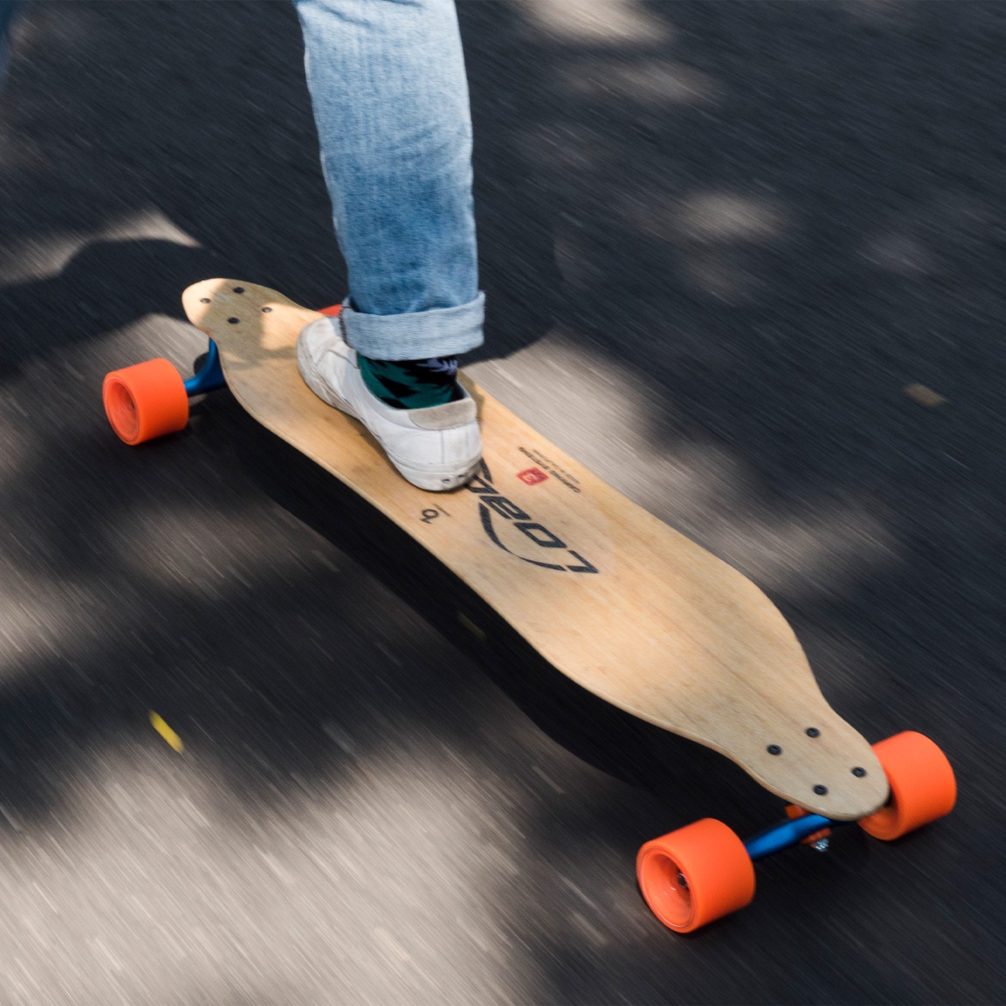 Rider's foot on a drop-through longboard with orange Orangatang In Heat 75mm wheels, motion-blurred pavement