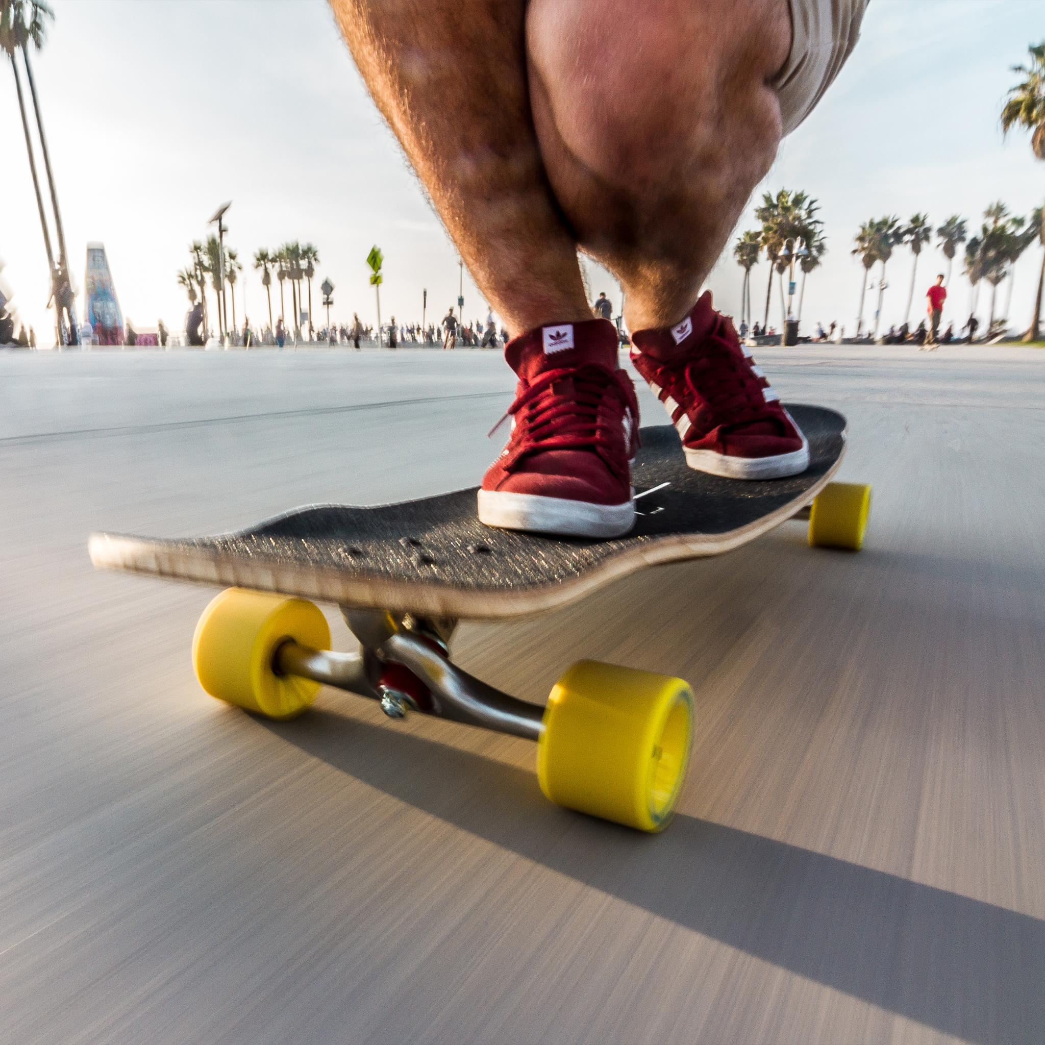 Skater in red sneakers pushing a board with yellow Orangatang Stimulus 70mm wheels along a palm-lined beach path