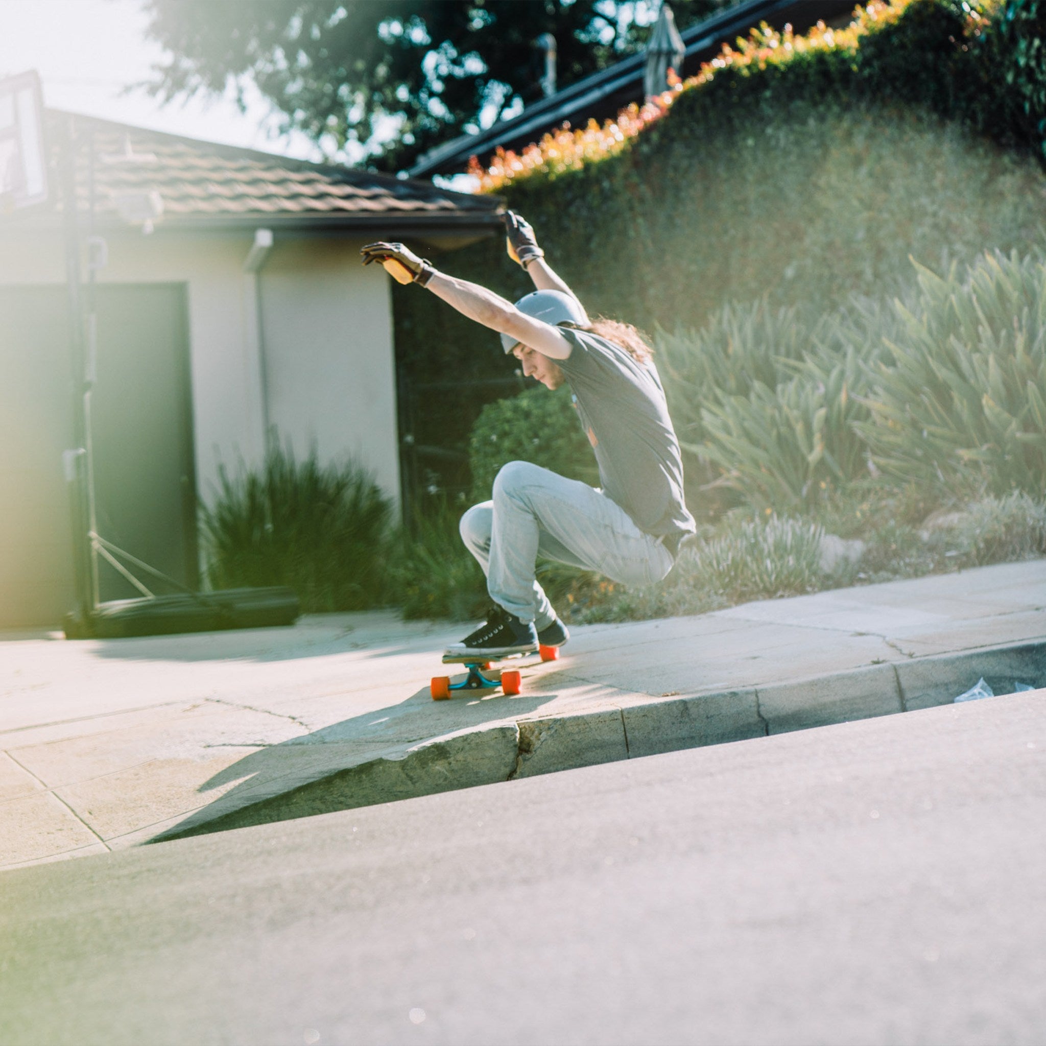 Skater ollieing a longboard with orange Orangatang Stimulus 70mm wheels off a suburban curb in bright sunlight