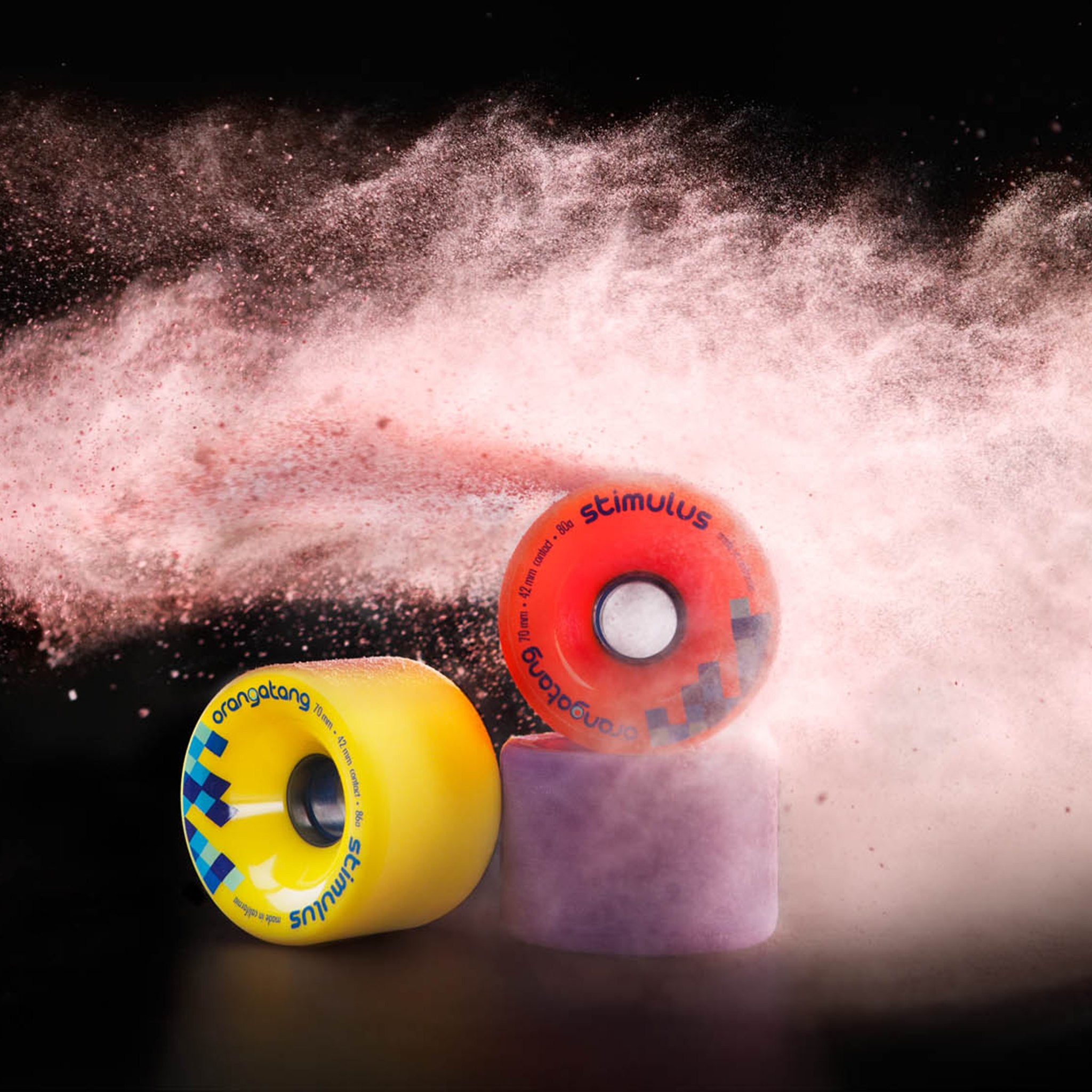 Orangatang Stimulus 70mm wheels in yellow, orange, and purple with a burst of pink powder on a black background